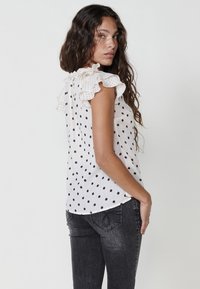 Blusa bianca senza maniche con pois neri, dotata di colletto arricciato e vestibilità ampia. Indossata con jeans scuri.