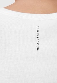 T-shirt bianco a coste con scollatura rotonda, con il nome del marchio "AULLSAINTS" e un logo nero di una capra ricamato sul retro.