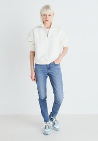 Fiatal, rövid szőke hajú személy fehér steppelt pulóverben, világoskék skinny farmernadrágban és kék-fehér sportcipőben áll egy sima fal előtt.