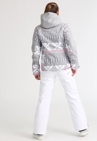 Grau gemusterte Snowboardjacke mit geometrischen Designs in Schwarz und Pink. Kombiniert mit weißen Schneehosen und grauem Schuhwerk.