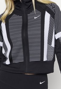 Chaqueta negra con cremallera y patrones de rayas geométricas blancas. Presenta un cuello alto y un dobladillo acanalado. El material parece elástico y deportivo.