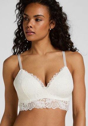 Frau mit lockigem Haar, die ein weißes Spitzen-Bralette mit gewellten Kanten und verstellbaren dünnen Trägern trägt und leicht zur Seite schaut.