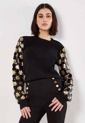 Mujer vestida con una blusa negra con mangas florales de lentejuelas doradas y negras y pantalones negros con botones dorados, de pie contra una pared blanca.
