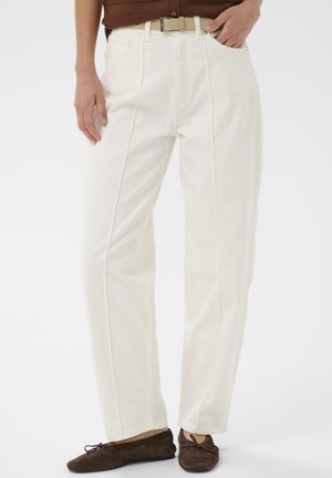 Weiße weite Hosen aus einem denimähnlichen Material, mit vorderen Taschen und einem sichtbaren Reißverschluss. Kombiniert mit braunem Schuhwerk.