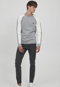 Grå sweatshirt med hvide raglanærmer, ribbede mansjetter og et lille sort logo på brystet. Sammen med mørke bukser og hvide sko.