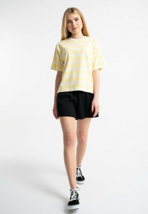 Młoda kobieta idąca, ubrana w żółtą pasiastą koszulkę, czarne szorty i czarne sneakersy za kostkę na białym tle.