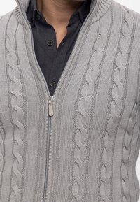 Lysegrå cardigan med lynlås og kabelstrikmønster, med høj krave og glat tekstur, lagdelt over en mørk skjorte med knapper.