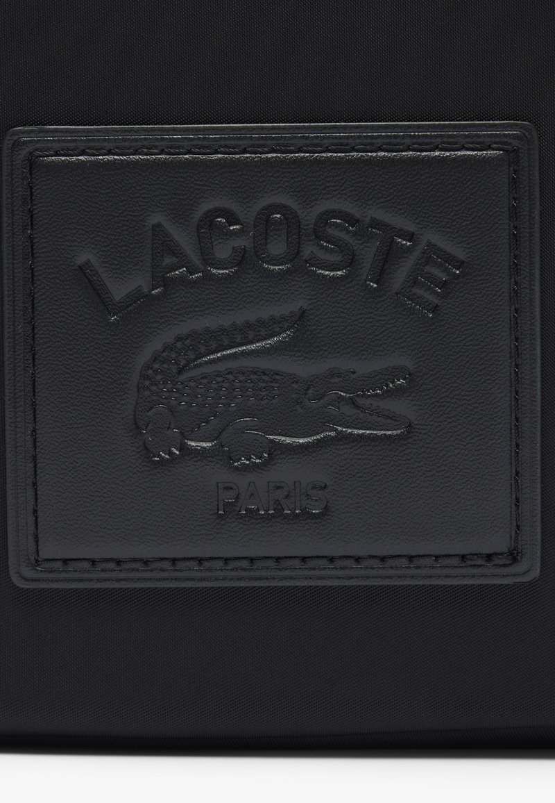 Patch Lacoste noire avec un logo crocodile texturé et une inscription en relief. Le patch est rectangulaire avec des bords cousus sur un fond en tissu.
