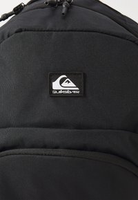 Mochila negra hecha de tejido duradero con una superficie texturizada, un bolsillo frontal y un parche del logo de Quiksilver. Tiene tiradores en los cierres.