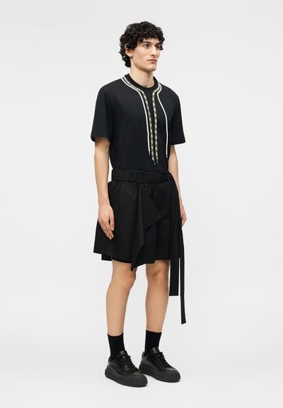 Mladý muž stojí a má na sobě černý krátký rukáv s vzorovanými kravatami, černou sukni s páskem, černé ponožky a černé tenisky.