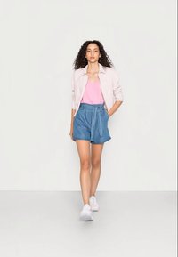 Jaqueta rosa clara sobre um top rosa em V, combinada com calções de ganga azuis atados na cintura. Modelo usa ténis brancos e tem cabelo encaracolado.