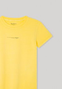 Camiseta amarilla de manga corta hecha de algodón suave. Presenta un cuello redondo y un sutil texto de la marca impreso cerca del cuello. Textura lisa.