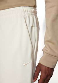 Nike sweatpants in light beige fabric, featuring an elastic waistband, side pockets, and a small, embroidered logo on the thigh.
