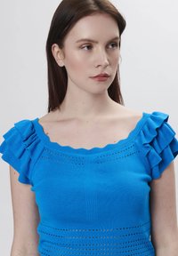 Royalblaue Strickoberteil mit rüschierten Ärmeln, mit einem gewellten Halsausschnitt und horizontalen Linienakzenten an der Taille.
