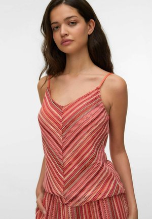 Kvinde med langt mørkt hår iført en ærmeløs, stribet rød og pink striktop og matchende bukser, stående mod en ensfarvet hvid baggrund.