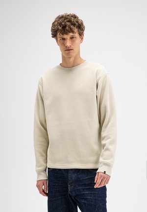 Jonge man met krullend haar die een beige, gestructureerd shirt met lange mouwen en donkerblauwe spijkerbroek draagt, staand tegen een effen lichte achtergrond.