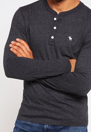 Mann trägt ein dunkelgraues Langarm-Henley-Shirt mit drei weißen Knöpfen und kleinem weißem Elch-Logo, die Arme vor der Brust verschränkt.