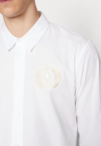 Camicia bianca a maniche lunghe in cotone. Presenta un logo rotondo ricamato in oro sul lato sinistro, colletto classico e una texture liscia.