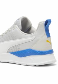 Grauer Sportschuh mit einem Obermaterial aus Netzgewebe, weißen Akzenten und einer blauen strukturierten Ferse. Mit einem gelben Logo nahe der Rückseite und einer flachen weißen Sohle.