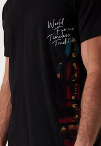 Sort bomulds T-shirt med korte ærmer. Har hvidt kursiv tekst og stort flerfarvet grafisk tryk på siden. Blød tekstur.