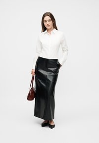 Γυναίκα που φοράει λευκό πουκάμισο με κουμπιά, μαύρη δερμάτινη μακριά φούστα, μαύρα μυτερά παπούτσια, κρατώντας καφέ τσάντα, στέκεται σε απλό λευκό φόντο.