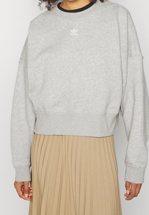 Kobieta ubrana w jasnoszary sweter Adidas z czarnym kołnierzem i beżową plisowaną spódnicę, pokazana od szyi do bioder.