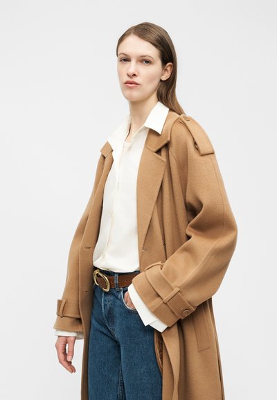 Donna con capelli castani lunghi che indossa un cappotto color cammello sopra una camicia bianca con colletto e jeans blu, in piedi con una mano in tasca.