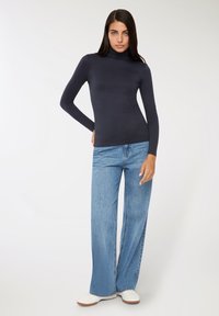 Kobieta ubrana w czarny sweter z długim rękawem, luźne jasnoniebieskie dżinsy i białe trampki, stojąca na tle czystej białej ściany.