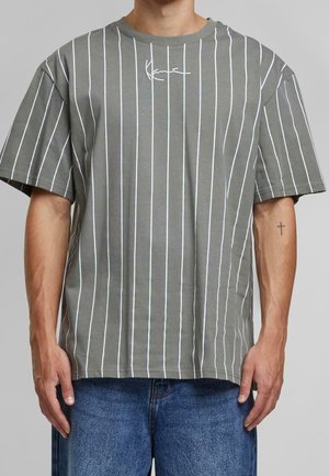 Man wearing gray T-shirt with vertical white stripes and small white logo on chest, paired with blue jeans against gray background.