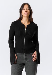 Pull noir côtelé avec un col rond, une fermeture éclair intégrale à l'avant et des manches évasées. Confectionné en tissu tricoté texturé, offrant une silhouette ajustée.