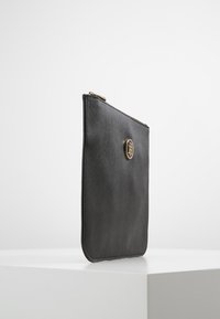 Clutch de cuero negro con una textura suave, que presenta un acento de logo en tono dorado y un cierre superior con cremallera. Forma rectangular y delgada.