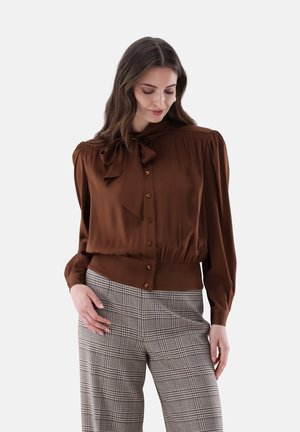 Frau trägt eine braune, geknöpfte Bluse mit einer Schleife am Hals und hoch taillierte karierte Hose, steht mit einem leichten Lächeln und schaut nach unten.