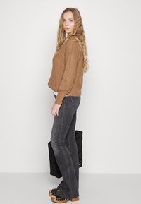 Lichtbruin gebreid sweater met ribbelstructuur, gecombineerd met donkergrijze flared jeans en zwarte platformklompen. Een zwarte gewatteerde tas wordt vastgehouden.