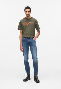 Olívazöld póló narancssárga logóval, kék, slim-fit farmernadrággal és fekete bokacsizmával párosítva. Egyszerű dizájn, minta vagy további részletek nélkül.