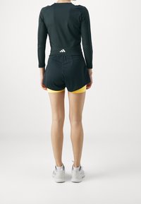 Svart långärmad topp med meshdetaljer, i kombination med svarta shorts som visar gula underbyxor. Vita sneakers fullbordar outfiten.