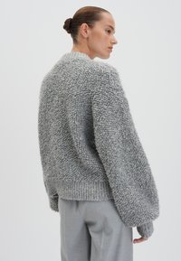 Maglione grigio chiaro con un motivo in maglia testurizzato e peloso. Presenta una vestibilità rilassata, colletto alto e orlo e polsini a coste. Indossato sopra pantaloni grigi chiari.