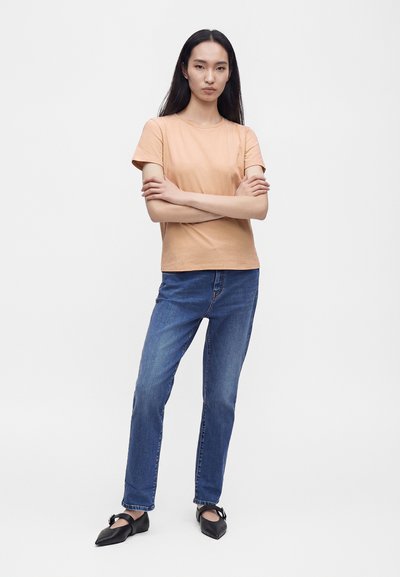 Femme aux longs cheveux noirs portant une chemise beige à manches courtes, un jean bleu et des ballerines noires pointues avec des brides, debout les bras croisés.