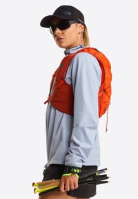 Pomarańczowa lekka kamizelka nawadniająca z regulowanymi paskami, noszona na jasnoniebieskiej bluzie z długim rękawem; trzyma kijki trekkingowe z zielonymi akcentami.