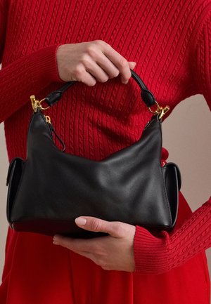 Mains tenant un sac à main en cuir noir avec des ferrures dorées, sur un fond de pull rouge en maille torsadée et de jupe rouge.