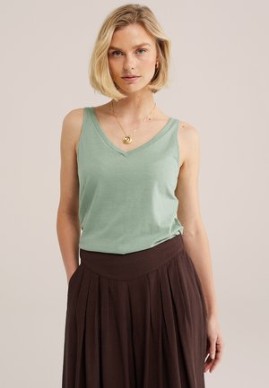 Mujer rubia con una blusa sin mangas de cuello en V verde claro, falda plisada marrón oscuro y collar dorado con un colgante en espiral.