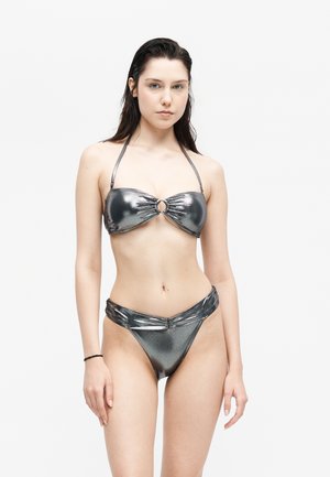 Nő áll, előre néz, fémes ezüst bikiniben, pántos felsőrésszel és magas kivágású alsóval, egyszínű fehér háttér előtt.