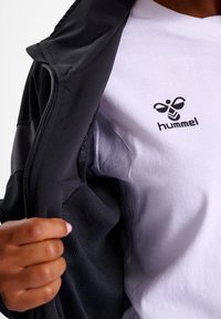 Svart zip-up jacka med texturerade detaljer, bärs över en vit t-shirt med en svart logotyp. Fokusera på dragkedjan och tygdetaljer.