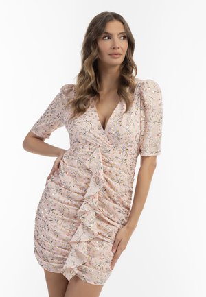Frau mit langen welligen Haaren, die ein rosa geblümtes, gerafftes Kleid mit Rüschen-Detail trägt und mit einer Hand auf der Hüfte zur Seite schaut.