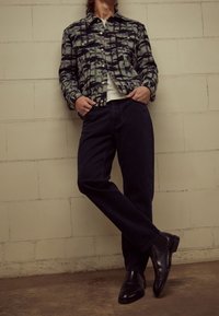 Rutig jacka i blå och gröna nyanser, vit t-shirt, mörk denimjeans och svarta läderskor, bärs av en person som lutar sig mot en vägg.