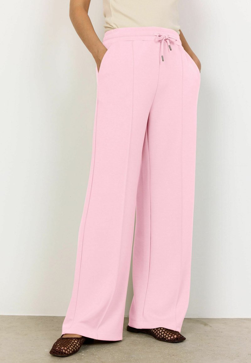 Persona con pantalones rosa de talle alto y pierna ancha, con cintura de cordón y bolsillos laterales, combinados con zapatos planos marrones tejidos.