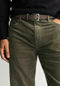 Olivgrüne Cordhosen mit einem strukturierten Rippmuster, ausgestattet mit Seitentaschen und einem dunkelbraunen Ledergürtel mit einer silbernen Schnalle.