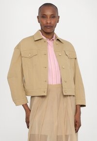 Khaki-denim takki, jossa on lyhennetty malli, nappikiinnitys ja kaksi etutaskua, yhdistettynä vaaleanpunaiseen kauluspaitaan ja läpinäkyvään beigeen hameeseen.
