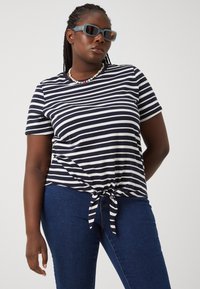 Camiseta a rayas en azul marino y blanco con mangas cortas y dobladillo anudado, combinada con jeans ajustados de color azul oscuro. Gafas de sol de color sólido y un collar de cuentas.