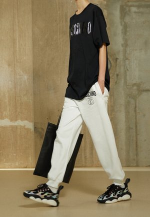 Osoba idąca, ubrana w czarny, oversize'owy T-shirt Moschino, białe spodnie dresowe Moschino, czarne buty sportowe w wzory i niosąca czarną torbę typu tote.