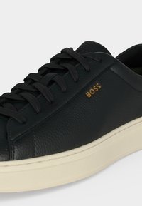 Basket en cuir noir avec lacets noirs, semelle blanche et logo doré "BOSS" sur le côté.